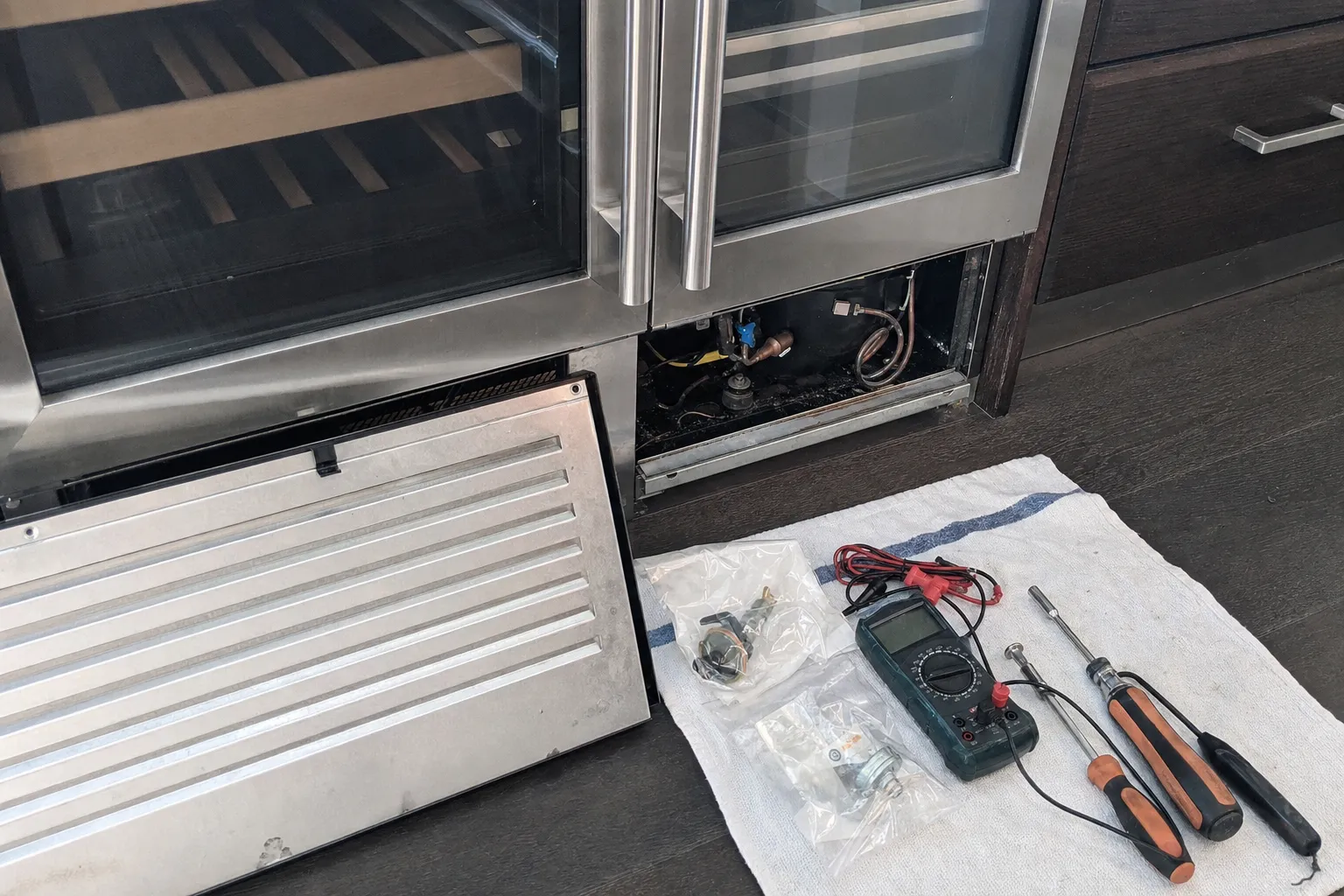 Base grille removed from a pair of undercounter glass-door wine coolers, tools and parts laid out on a cloth