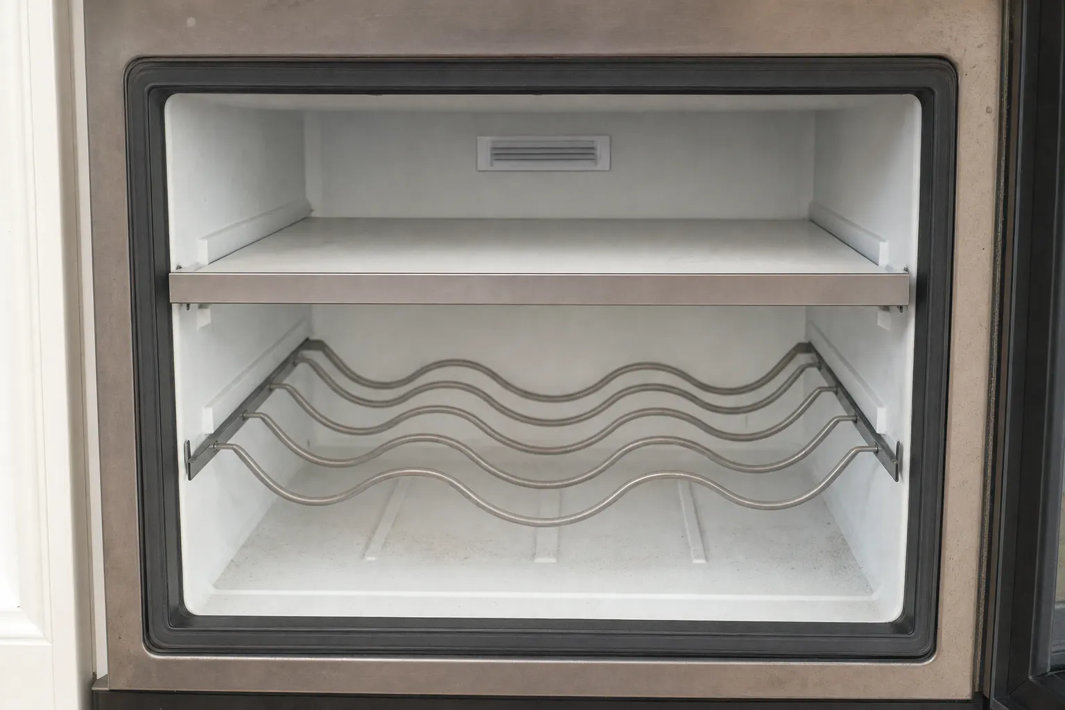 Open stainless undercounter compartment with a shelf above a wavy wire bottle rack, an air vent on the rear wall and a black gasket around the opening