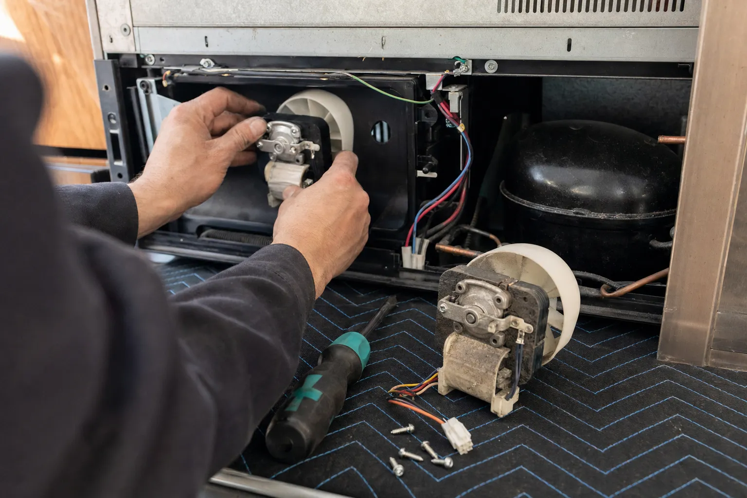 Hands fitting a fan motor with a white impeller into the machine compartment beside the compressor, the dusty old motor, a screwdriver and screws on a quilted blanket